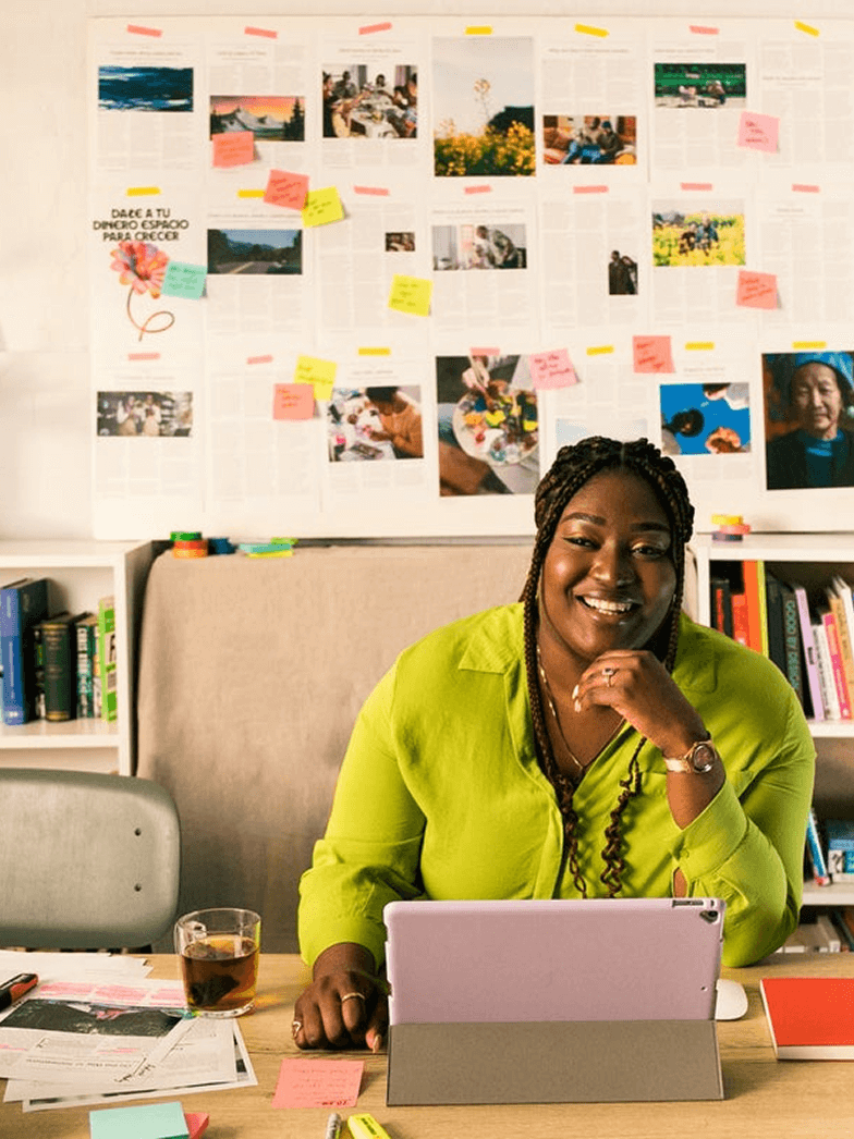 Una donna con una maglietta verde è seduta a una scrivania e sorride. Di fronte a lei c'è un tablet viola appoggiato su una custodia grigia. Dietro di lei, una parete è coperta da varie foto, disegni e numerosi post-it colorati, che suggeriscono uno spazio di lavoro creativo. Le parole "FOR STARTUPS" sono visualizzate in testo bianco nella parte superiore dell'immagine, e un cerchio verde con un segno più bianco si trova nell'angolo in basso a destra.